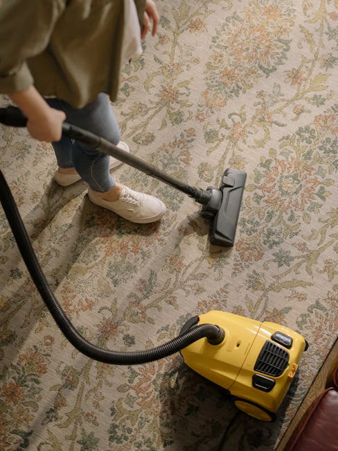 A professional cleaner dressed in a full white protective suit, face mask, and orange gloves is operating a vacuum cleaner in a modern, well-lit living room. The room features a sleek, gray-toned tile