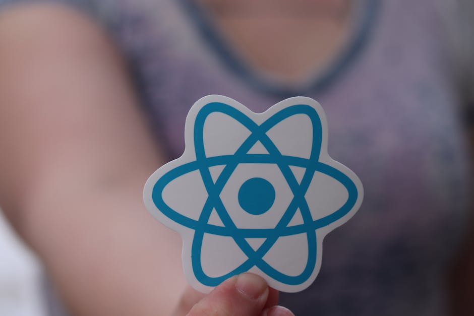 A person is holding a sticker with a blue atomic symbol featuring orbit lines and a central dot, positioned in front of their chest. The individual is wearing a short-sleeved shirt in a mottled purple